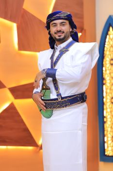 Young man in traditional clothing smiles indoors with decorative background.