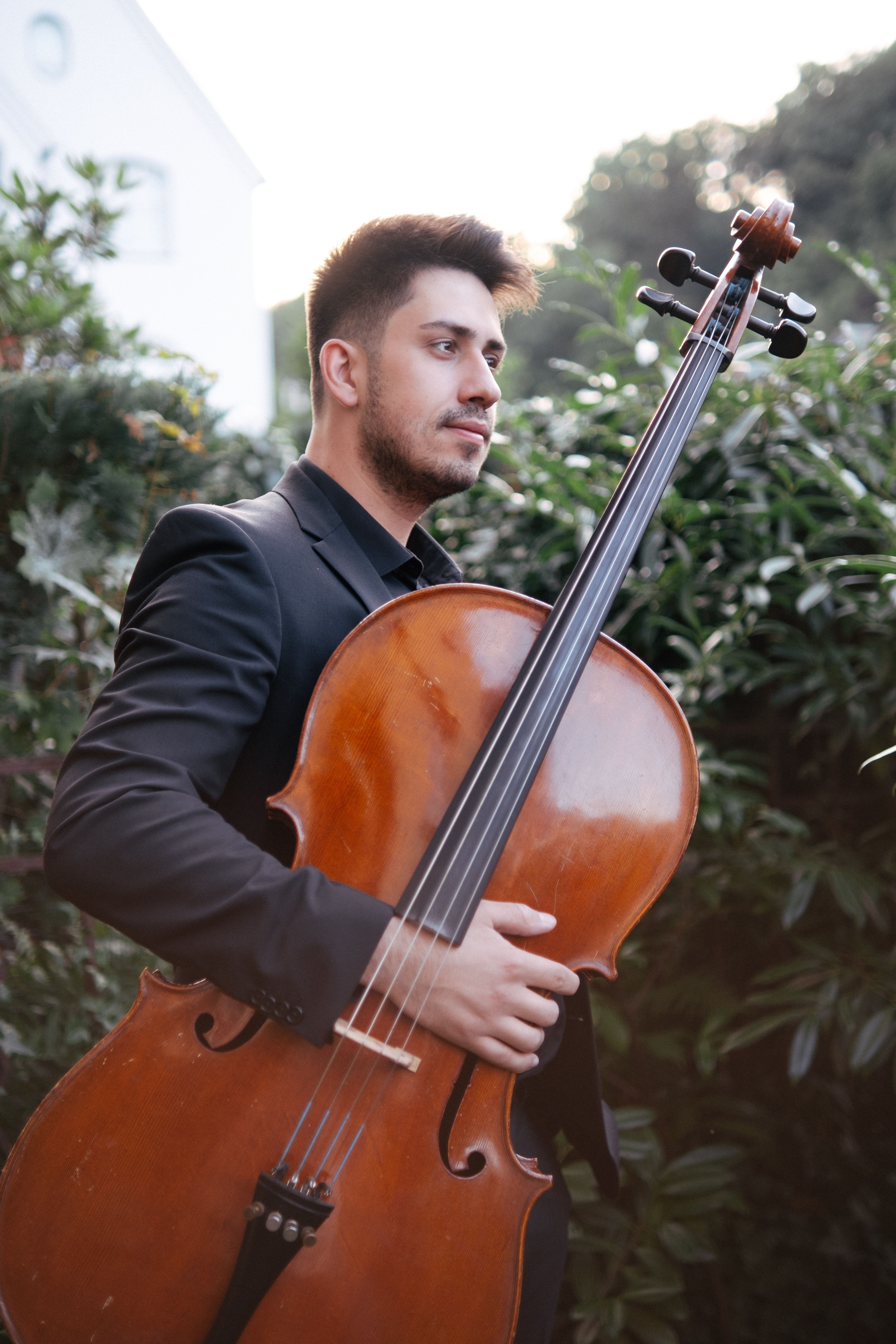 A young musician in a suit plays the cello outdoors, surrounded by lush greenery.