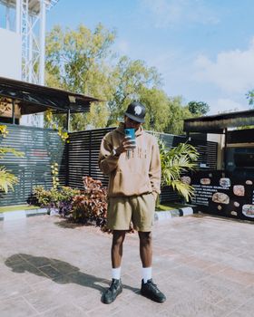 Young man in stylish urban outfit taking a selfie outdoors with foliage backdrop.