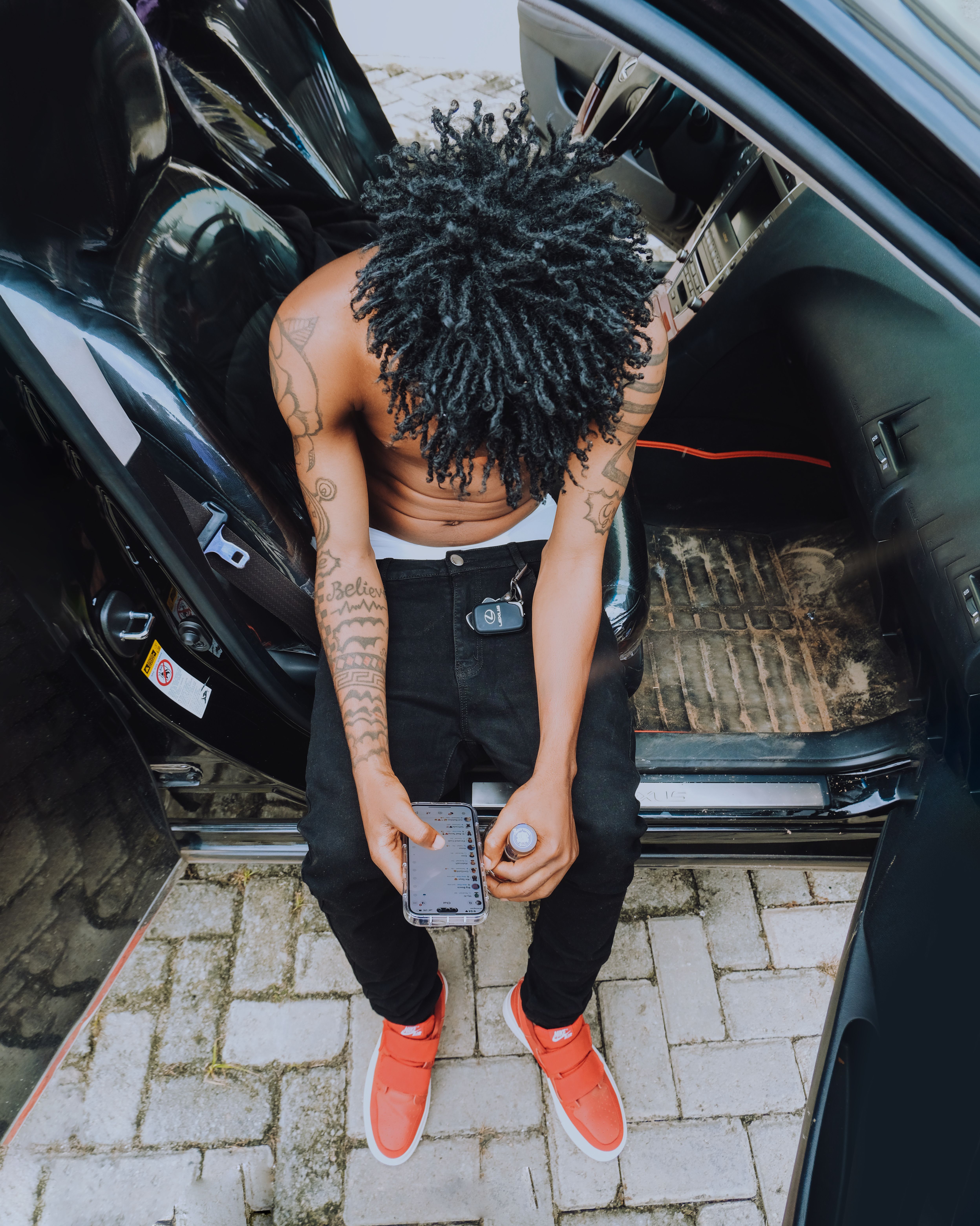 Young tattooed man looking at smartphone while sitting in car doorway, showcasing urban lifestyle.