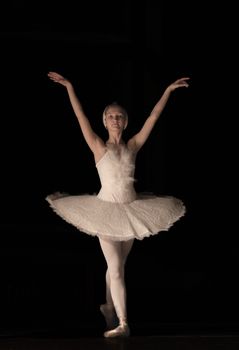 Ballet dancer in a white tutu gracefully poses on a dark stage.