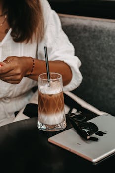 Woman enjoys iced coffee at a cozy café, tablet and sunglasses on table.