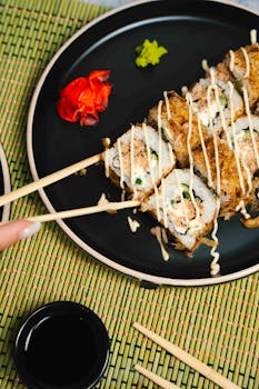 Close-up of sushi roll with chopsticks on a black plate, garnished with wasabi and ginger.