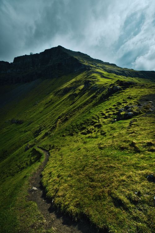 Free A scenic mountain path leading up a green hillside under a dramatic, cloudy sky. Stock Photo