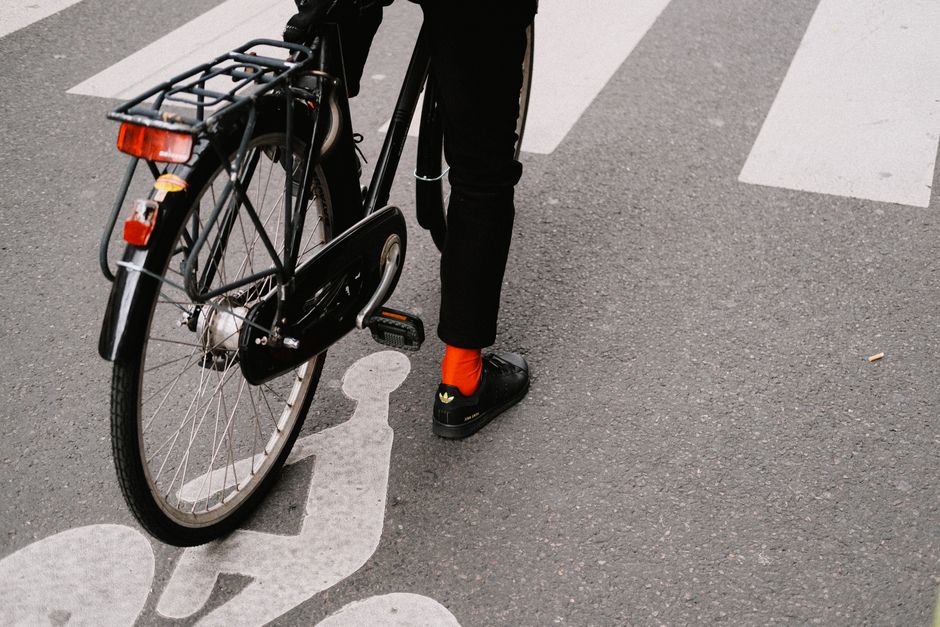 A cyclist wearing black pants and red socks stands with a bike on an urban street.
