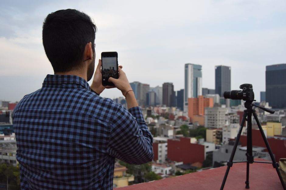 Man photographing city skyline with smartphone and camera on tripod from a rooftop.