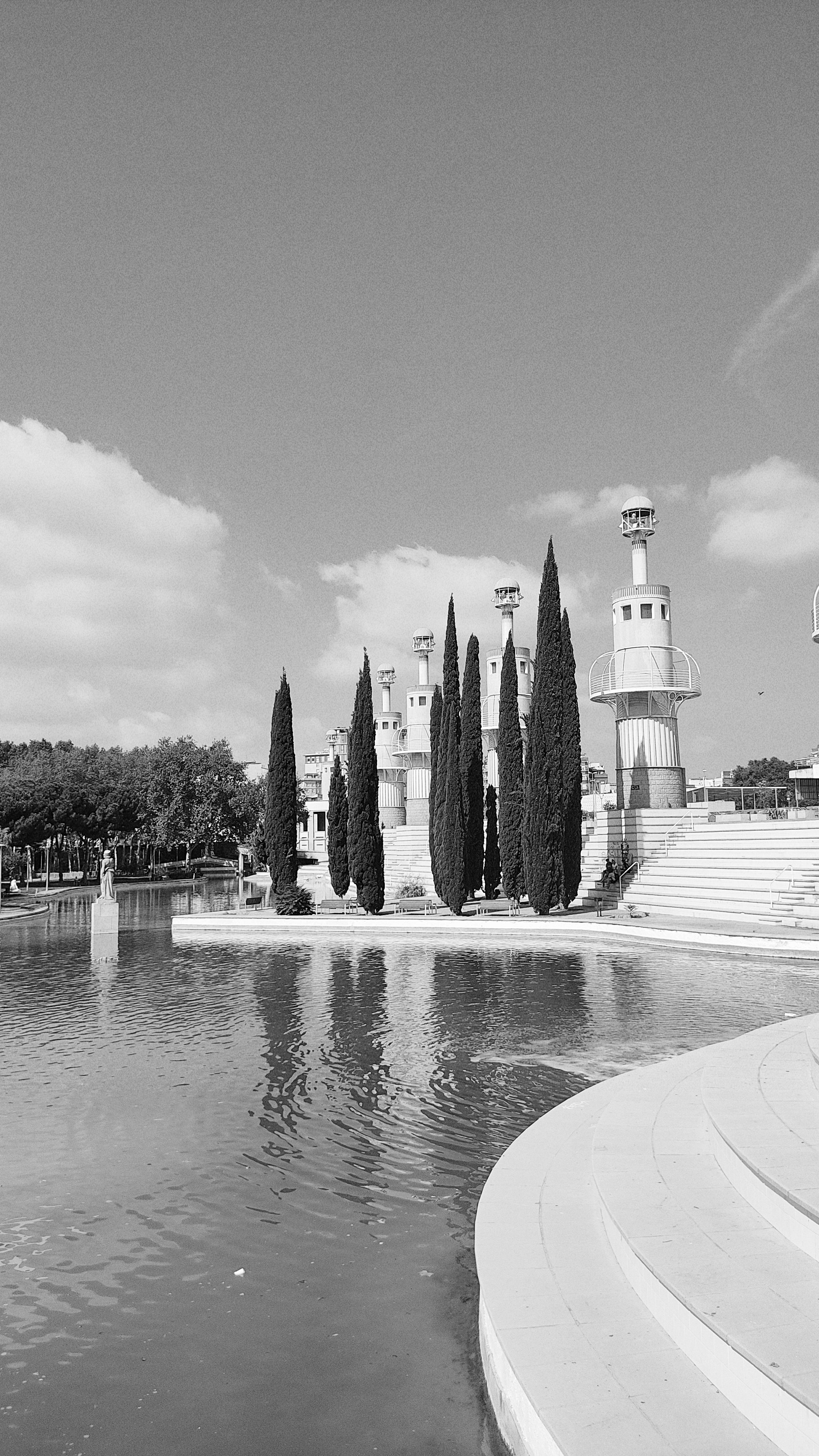 Black and white image of modern towers beside a tranquil park lake with clear skies.