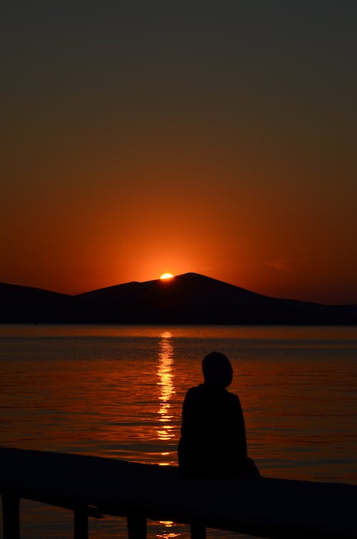 Silhouette of a Person Sitting on the Shore at Sunset 