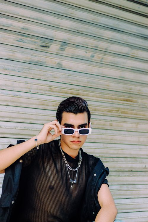 Free A fashionable young man wearing sunglasses and jewelry posing against a shuttered background outdoors. Stock Photo