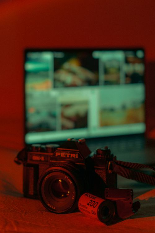 Free Close-up of a vintage camera and film roll in moody lighting with a blurred laptop screen in the background. Stock Photo