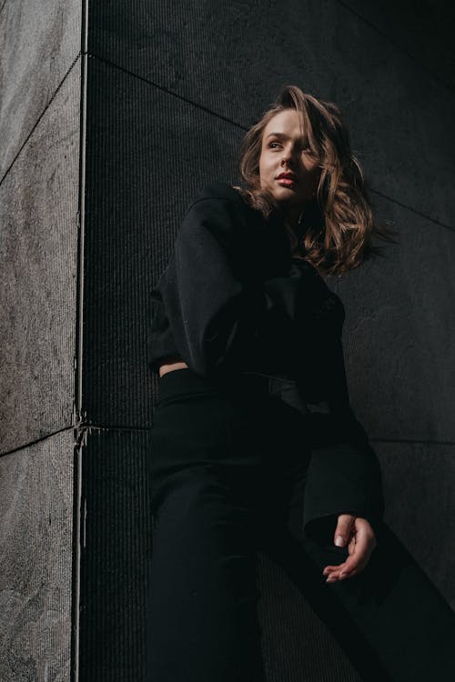 A woman in black pants and a black top leaning against a wall