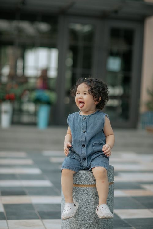 Free A young girl in overalls sits smiling on a stone pillar in a city setting. Stock Photo