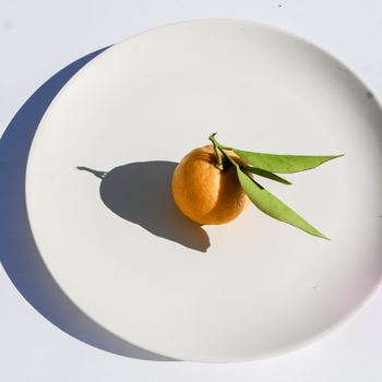 Minimalist image of a fresh orange with leaves on a white plate, ideal for food and health themes.