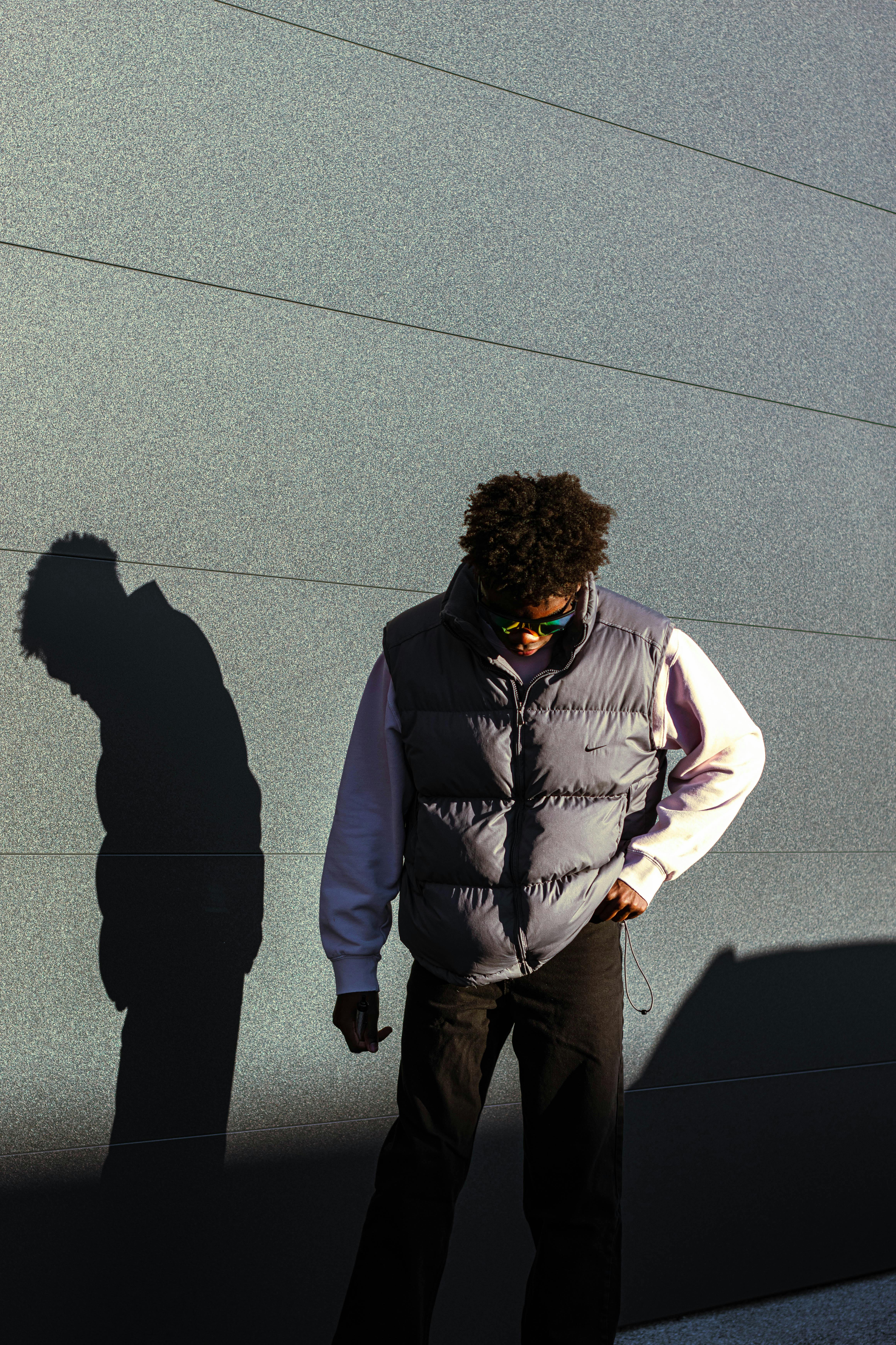 Free Stylish young man with afro hair in a puffer vest casts shadow on wall, showcasing urban fashion. Stock Photo