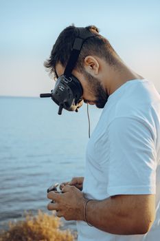 Young man using drone equipment with goggles on a coastal shore, showcasing technology in nature.