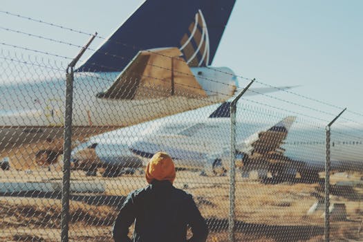 A person in a hoodie watches airplanes through a chain-link fence, capturing urban travel inspiration.