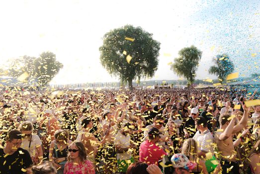 A lively crowd enjoying an outdoor music festival with confetti under a sunny sky.