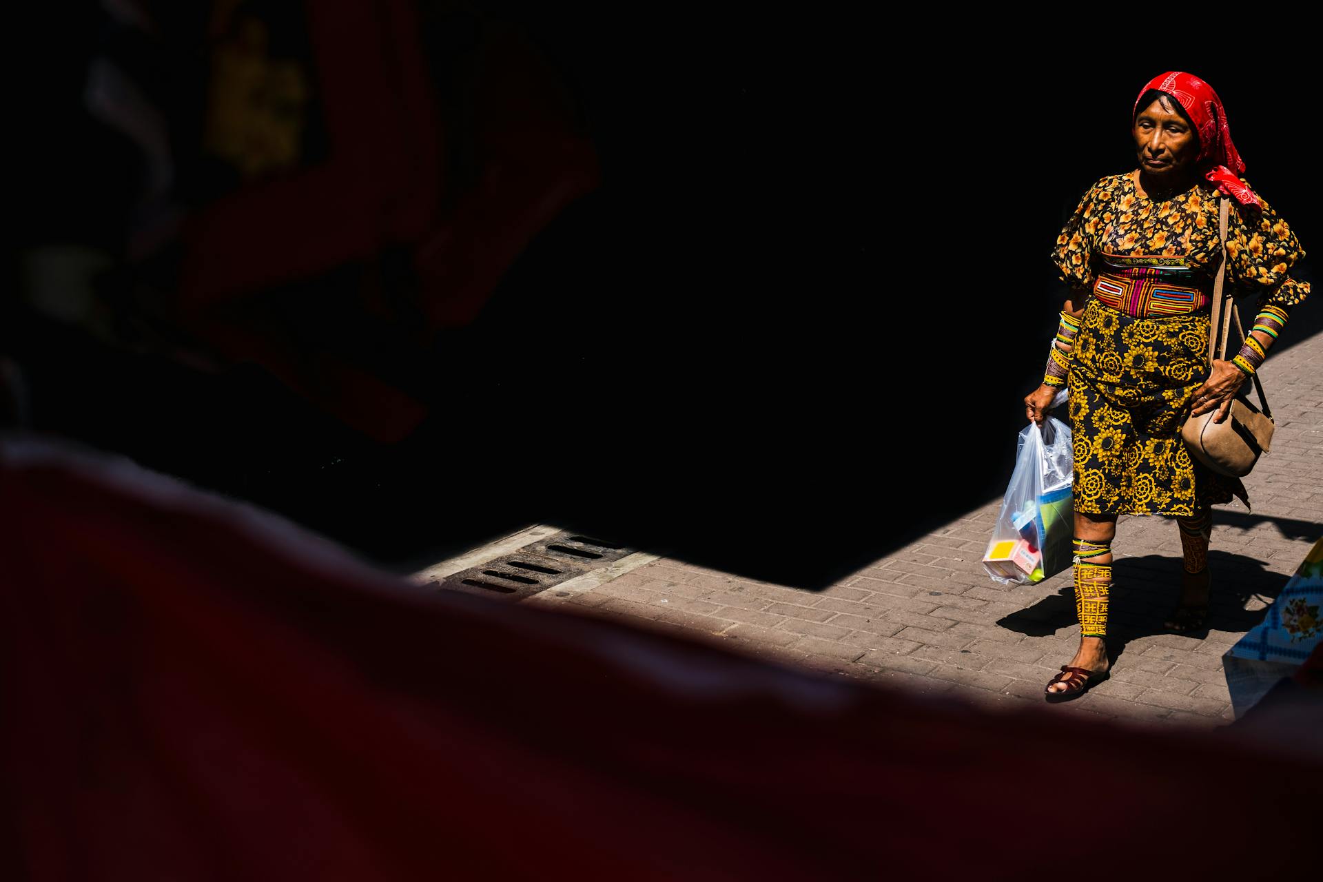 A woman wearing a vibrant traditional dress walks in Santa Ana, Panama, carrying shopping bags on a sunny day.