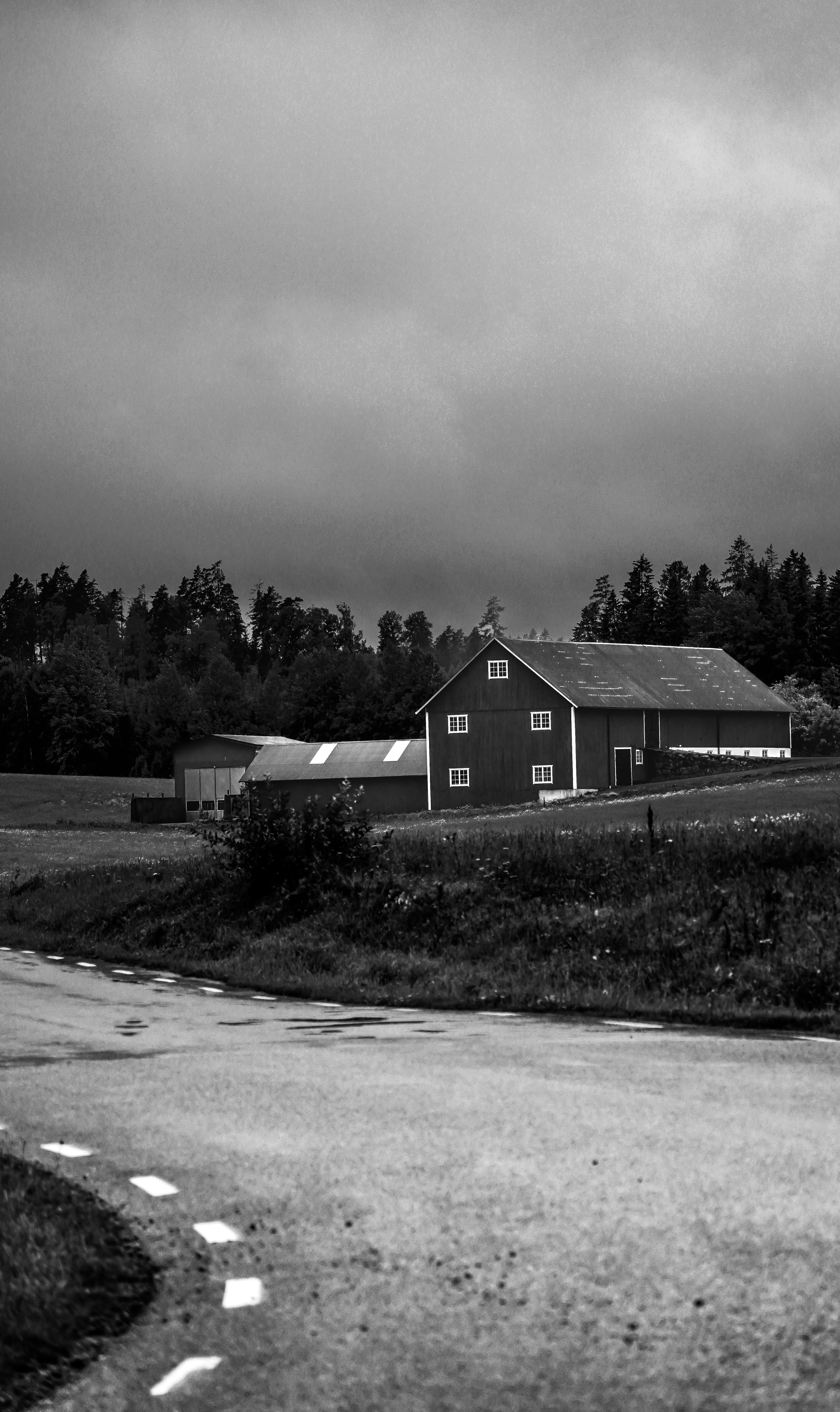 A black and white image of a rural farmhouse with a curving road and cloudy sky.