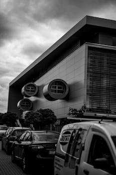 Black and white photo of modern urban architecture with parked cars beneath a cloudy sky.