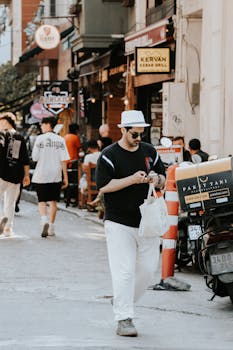 A fashionable man in casual attire walks down a bustling city street on a sunny day.