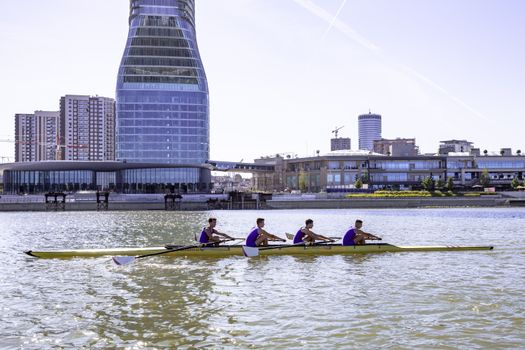 A rowing team practices on calm waters with a backdrop of modern skyscrapers.