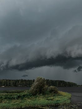 A powerful storm cloud formation looms over a serene lake landscape, showcasing extreme weather.