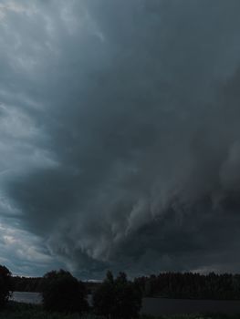 A dramatic storm cloud looms over a rural landscape with trees and a lake.