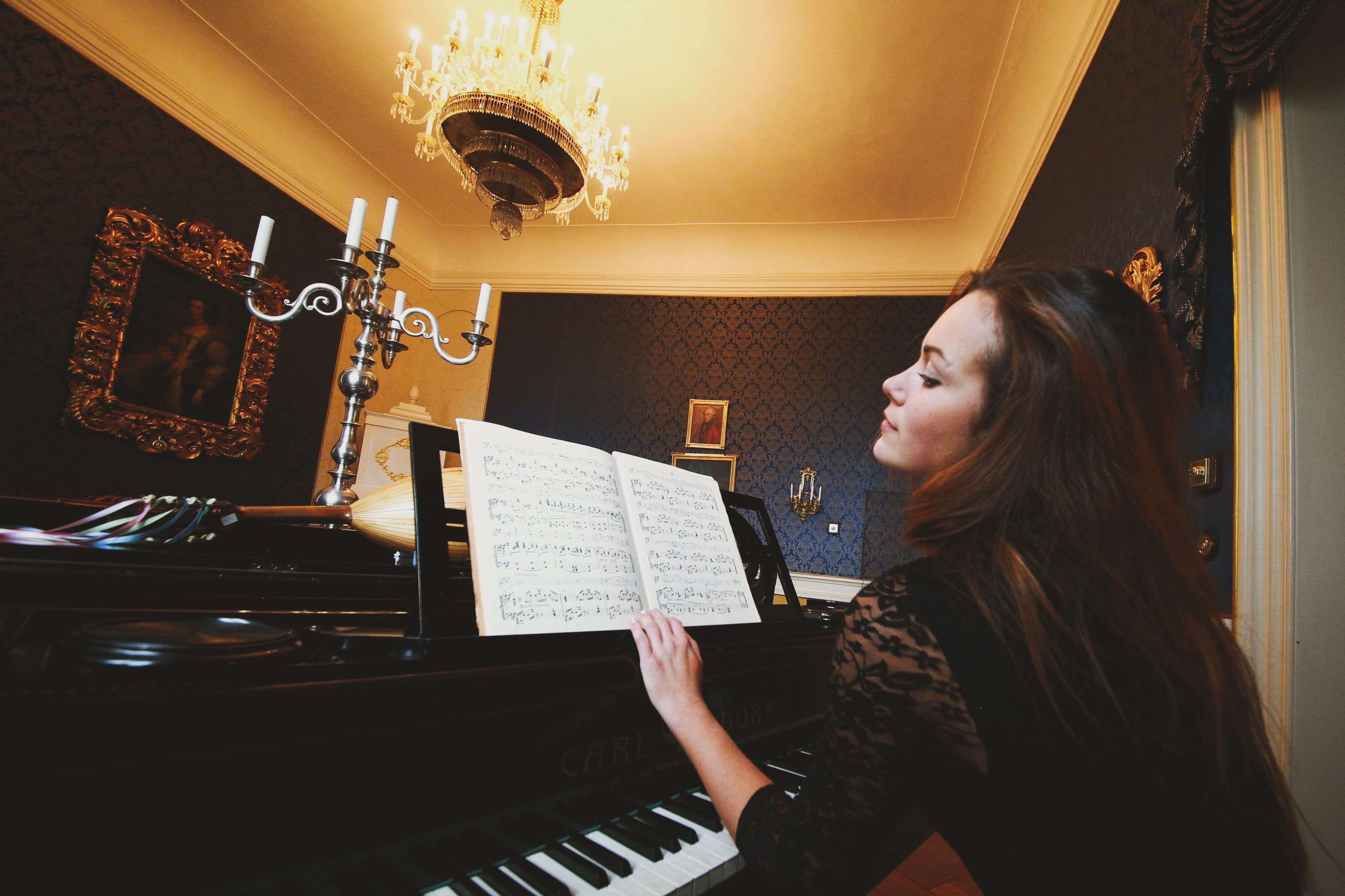 A woman with long hair plays piano under a chandelier in a classic, elegant room.