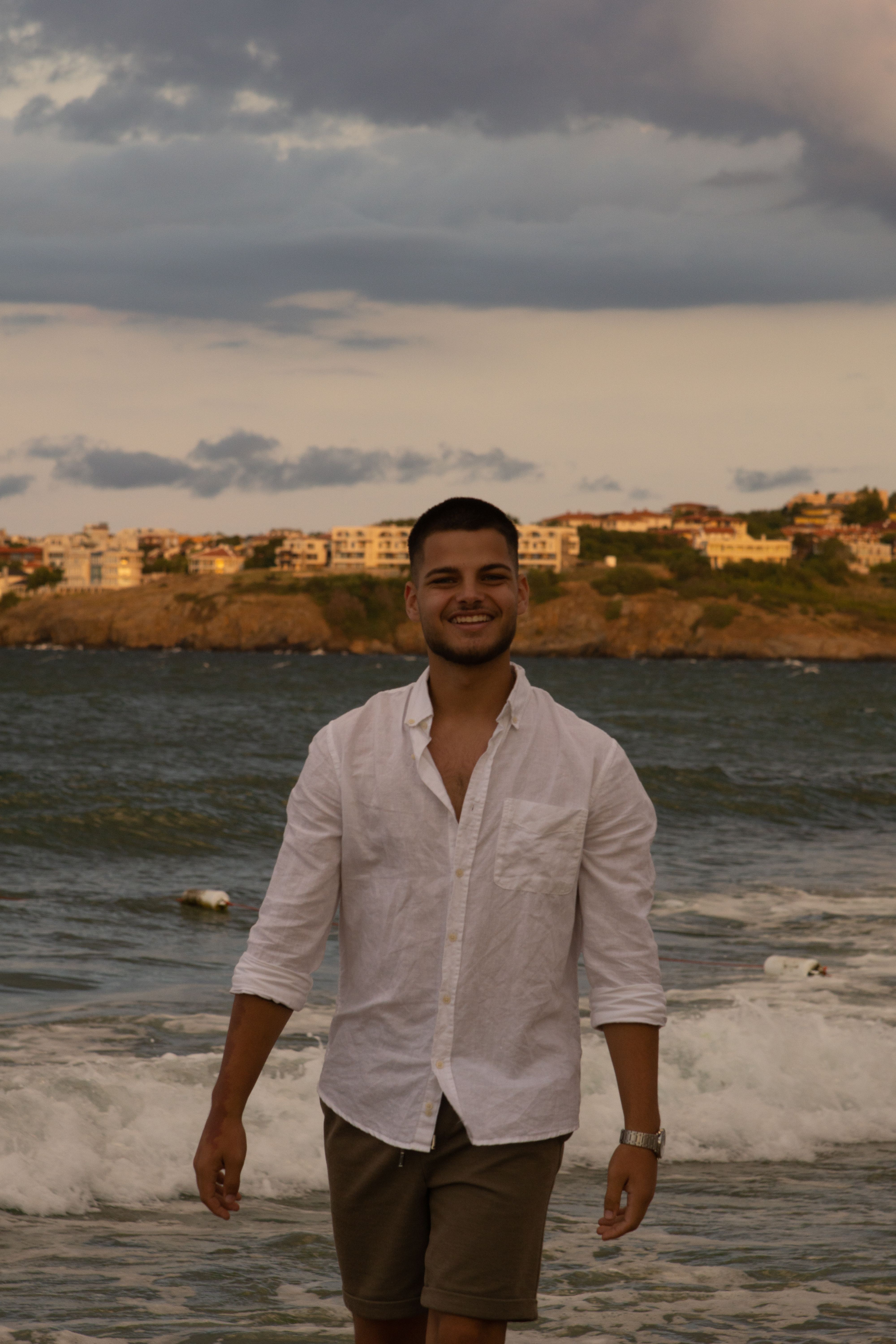 A man wearing a white shirt enjoys a walk along the seaside during sunset, exuding a relaxed vacation vibe.