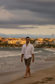 A young man leisurely walks along a picturesque beach at dusk, enjoying the serene atmosphere.