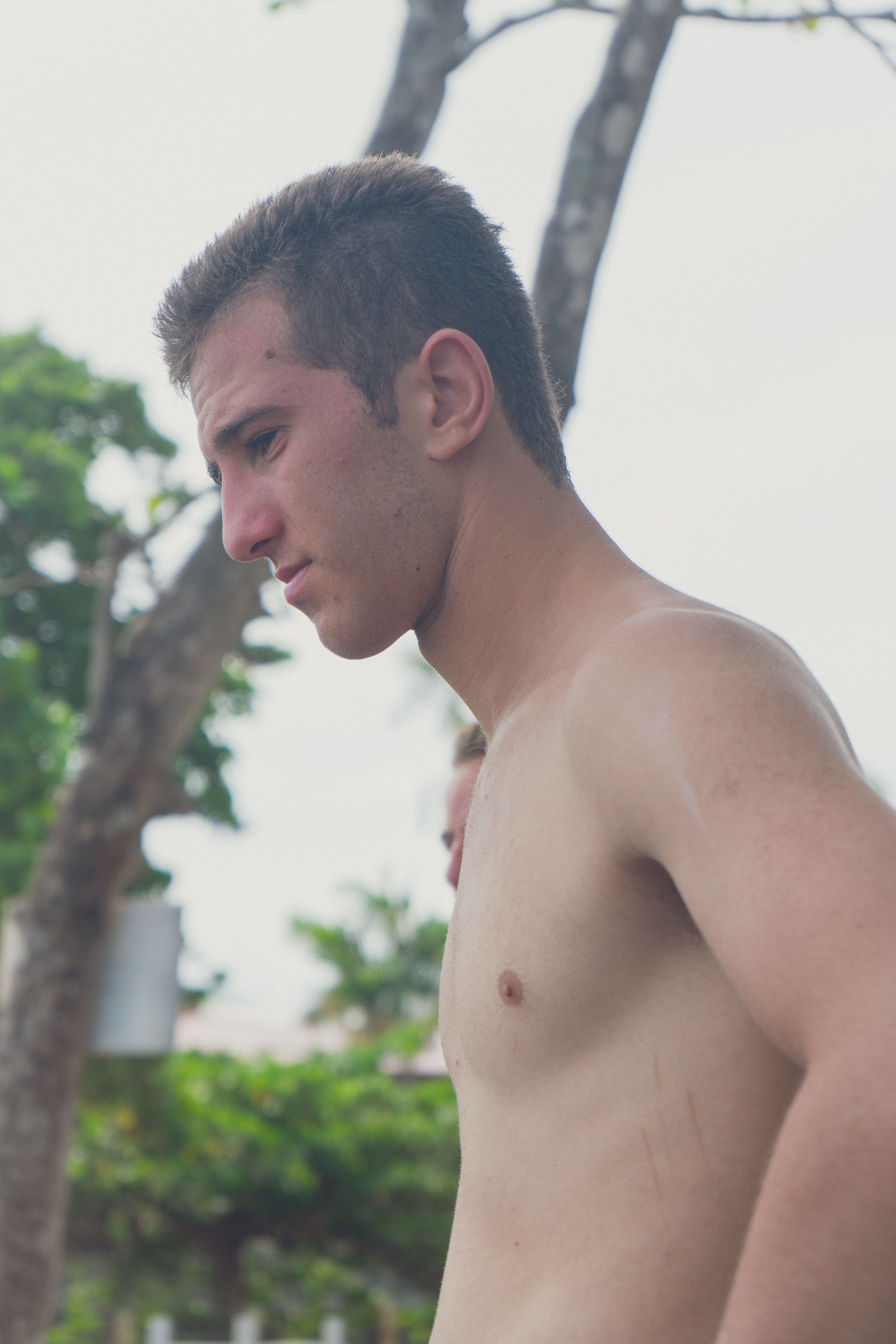 A young man with short hair looks thoughtful while standing outdoors in a lush, natural environment.