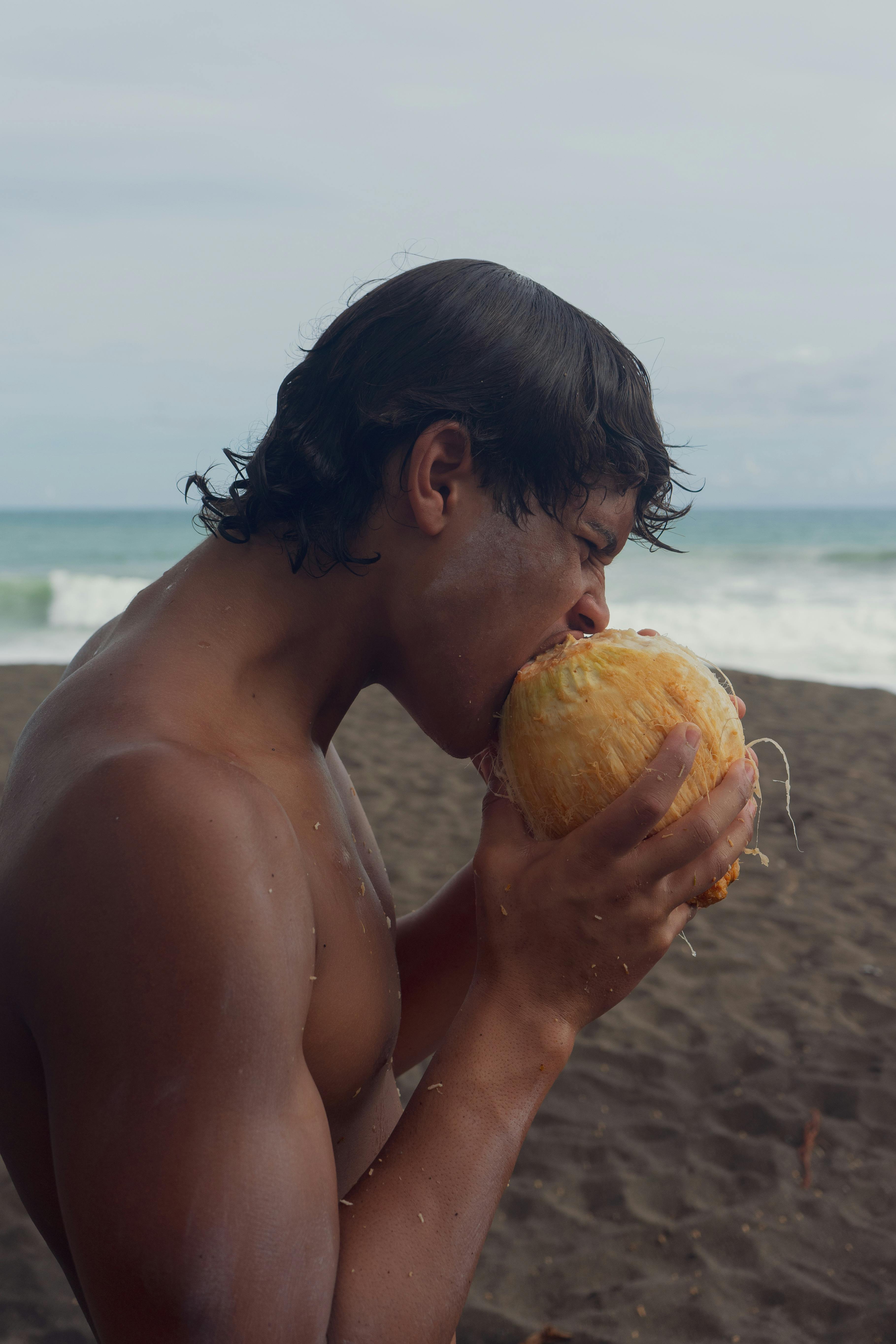 Topless man biting a coconut on the beach with the sea in the background.