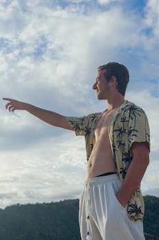 A smiling young man in a tropical shirt points towards the horizon against a cloudy sky.