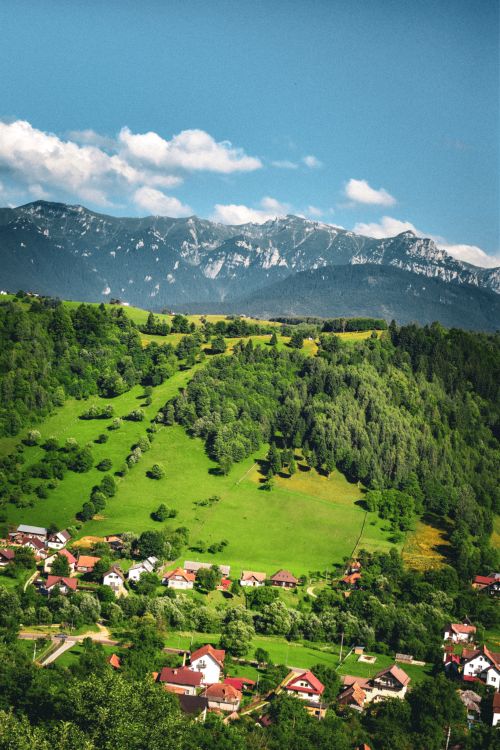Free Aerial view of a picturesque mountain village surrounded by vibrant green hills and majestic mountains under a blue sky. Stock Photo