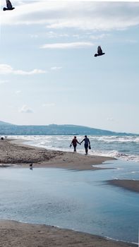 Couple holding hands on a sandy beach with waves and birds in flight, under a clear sky.