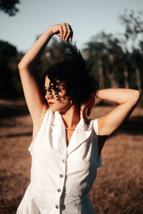 Free A woman with curly hair poses stylishly outdoors during a sunny summer evening. Stock Photo