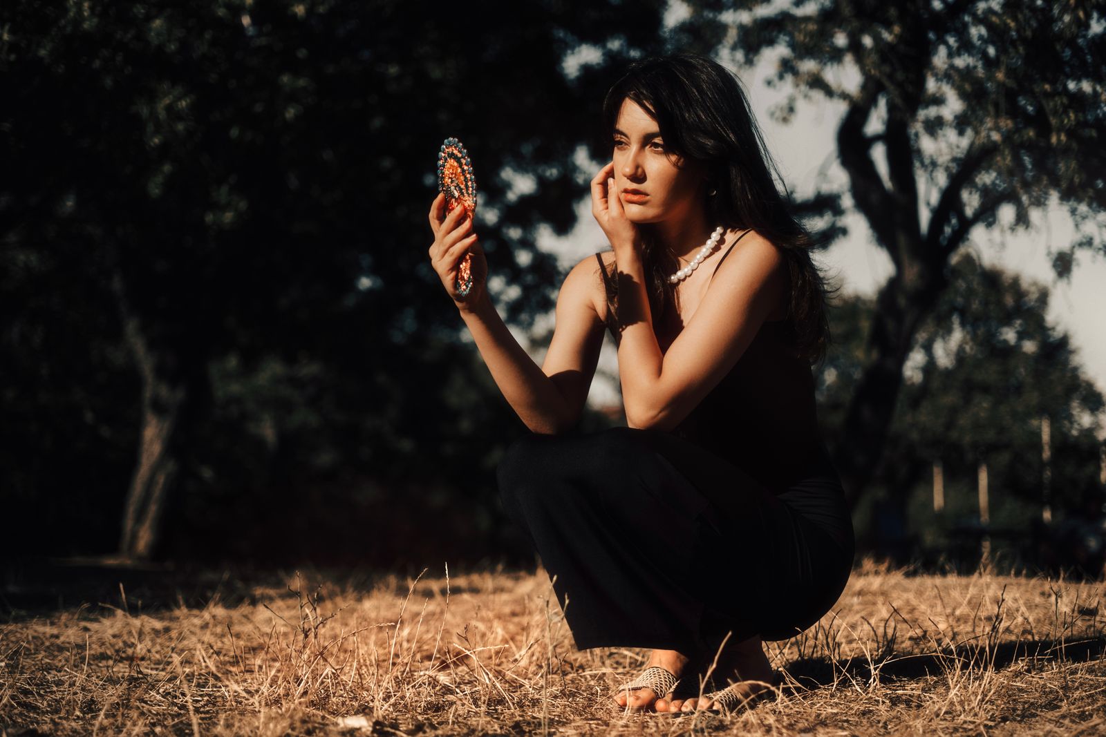Woman kneeling outdoors in golden hour light looking at her reflection in a small hand mirror.