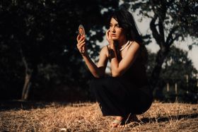 Woman kneeling outdoors in golden hour light looking at her reflection in a small hand mirror.