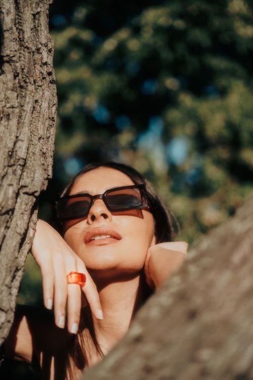 Free A fashionable woman wearing sunglasses enjoys a sunny day outdoors, framed by tree branches. Stock Photo