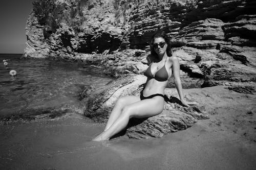 Woman in Bikini and Sunglasses Sitting on Rocks on Sea Shore