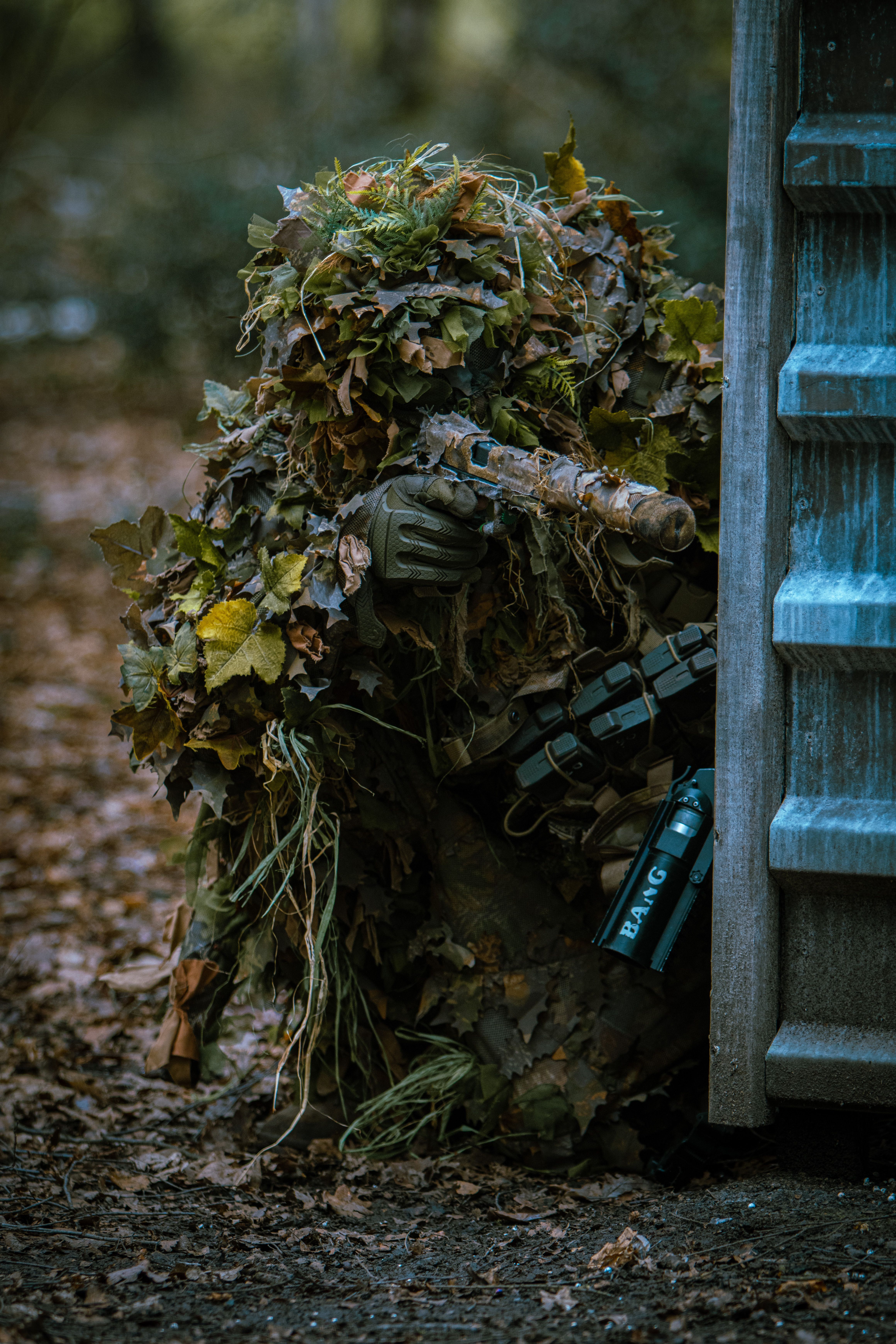 A soldier in full camouflage gear aiming a weapon in a forest, blending with foliage.