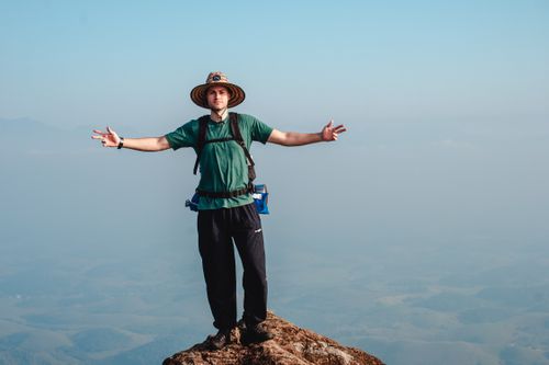 Free Man with arms outstretched stands on a mountain peak, enjoying scenic views. Stock Photo