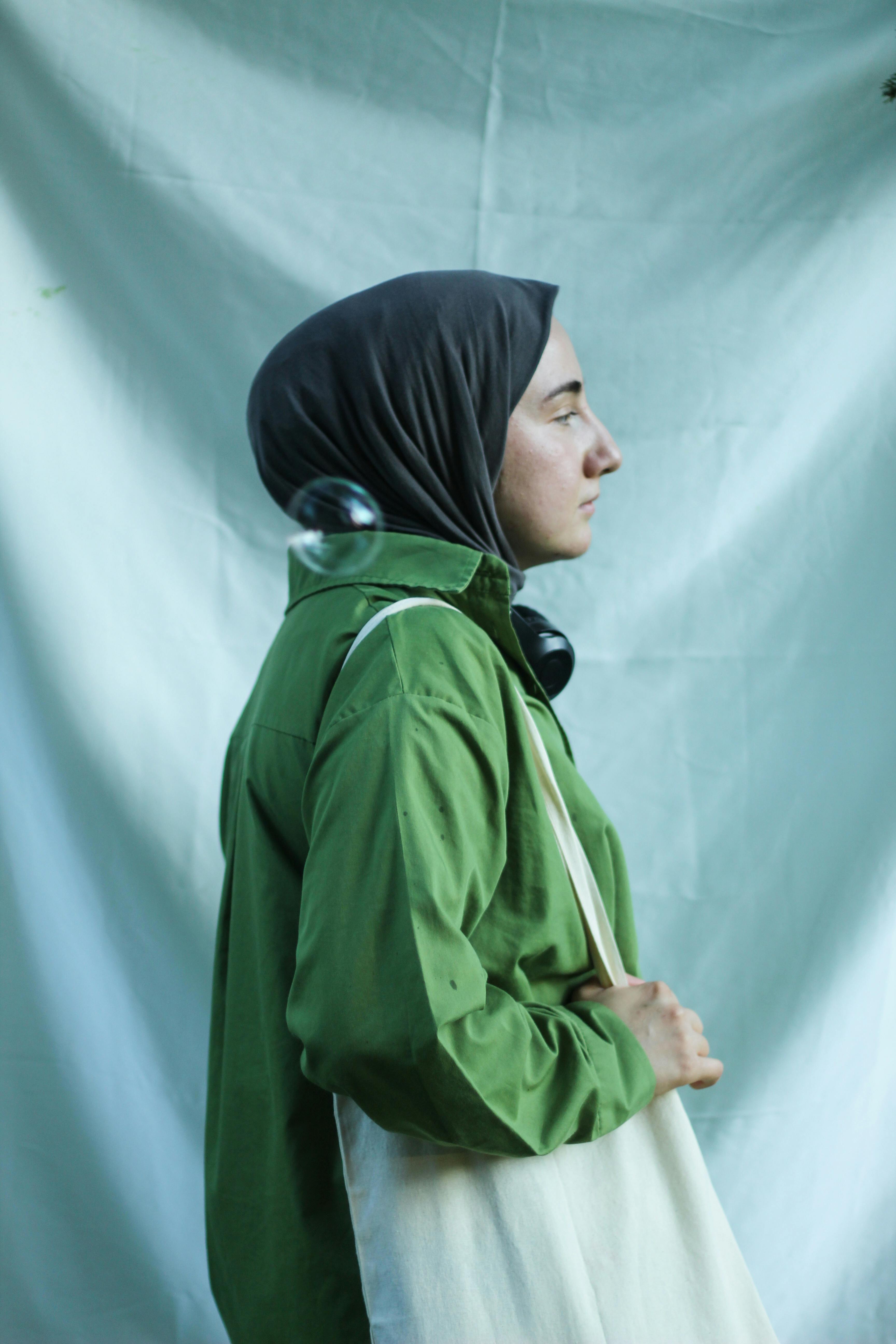 A woman in a green outfit and hijab holding a tote bag in profile view.