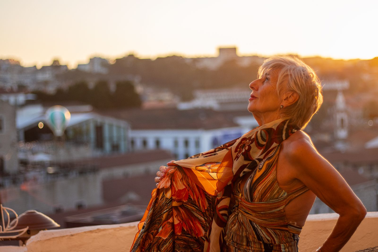 A senior woman elegantly poses in a colorful dress on a rooftop during a warm sunset in the city.