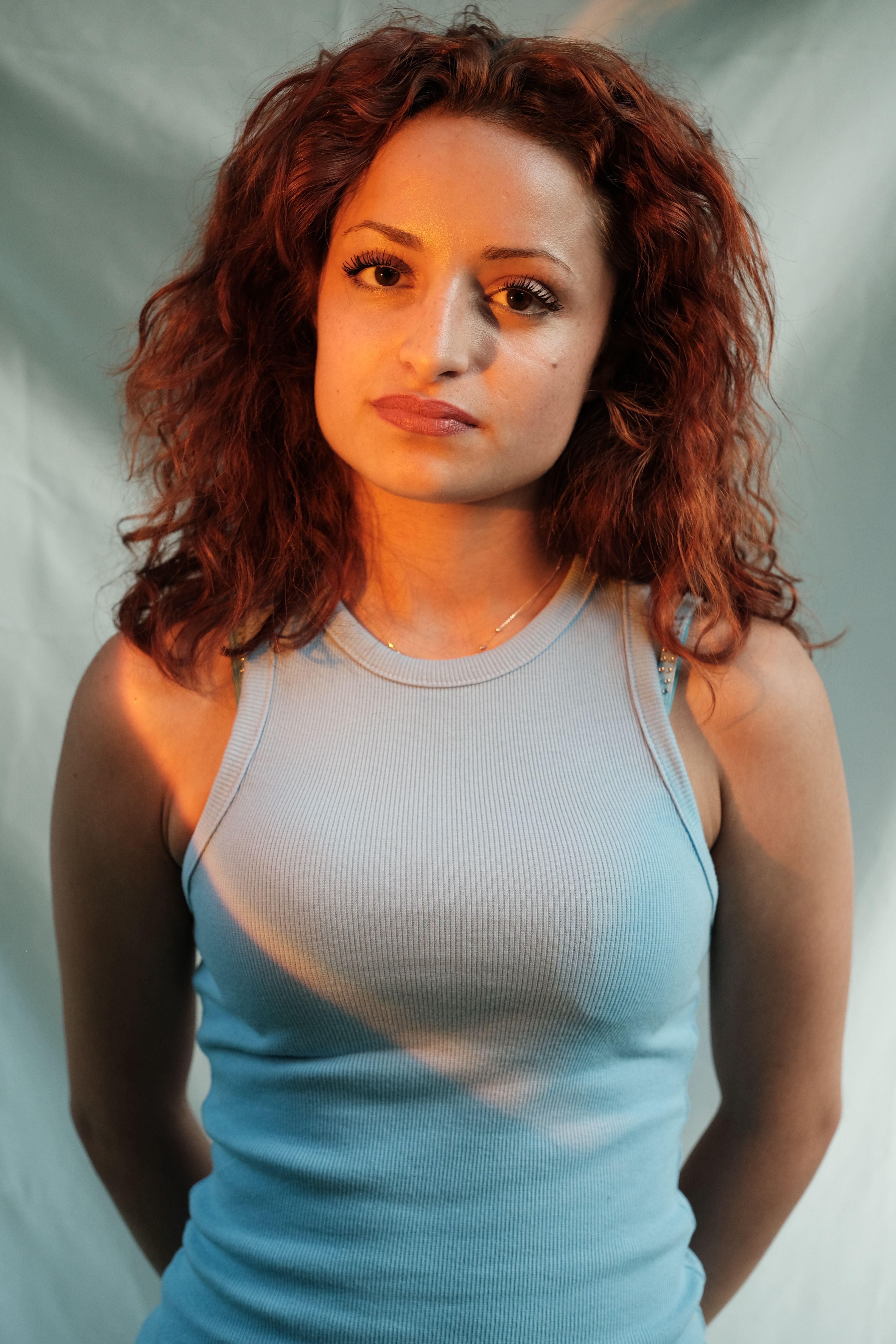 A young woman with curly hair wearing a light blue ribbed dress poses in a studio setting.