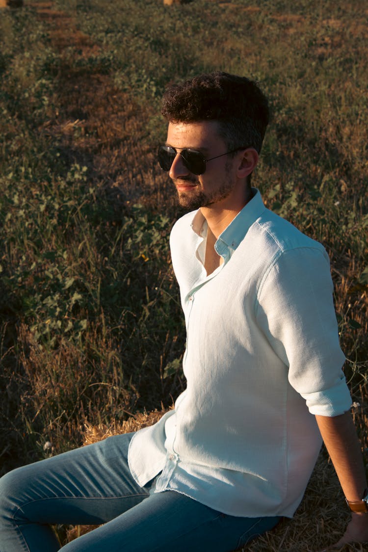 Brunette Man In White Shirt And Jeans Sitting On A Hayfield