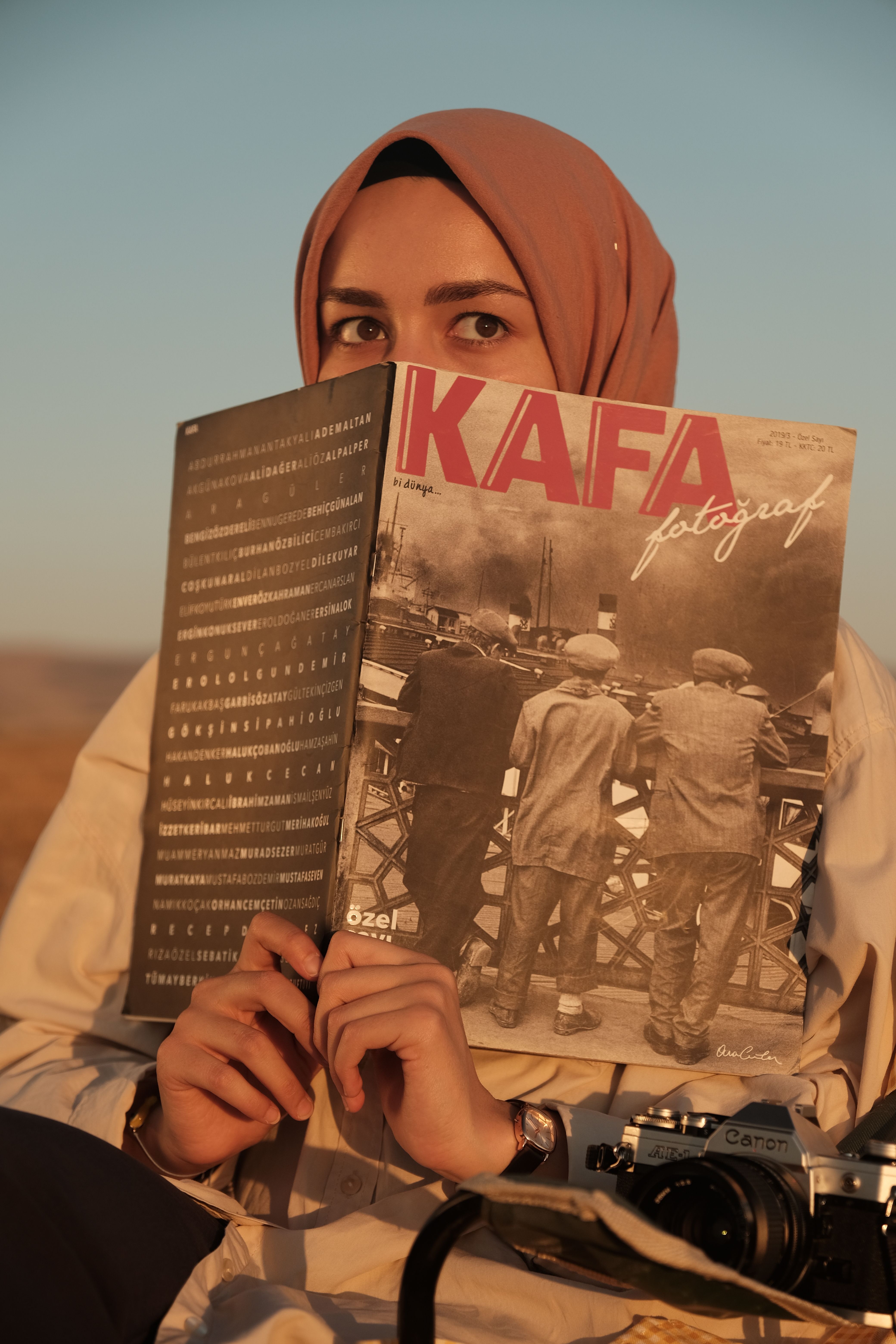 Woman in hijab reading a magazine outdoors during sunset, with a vintage camera beside her.