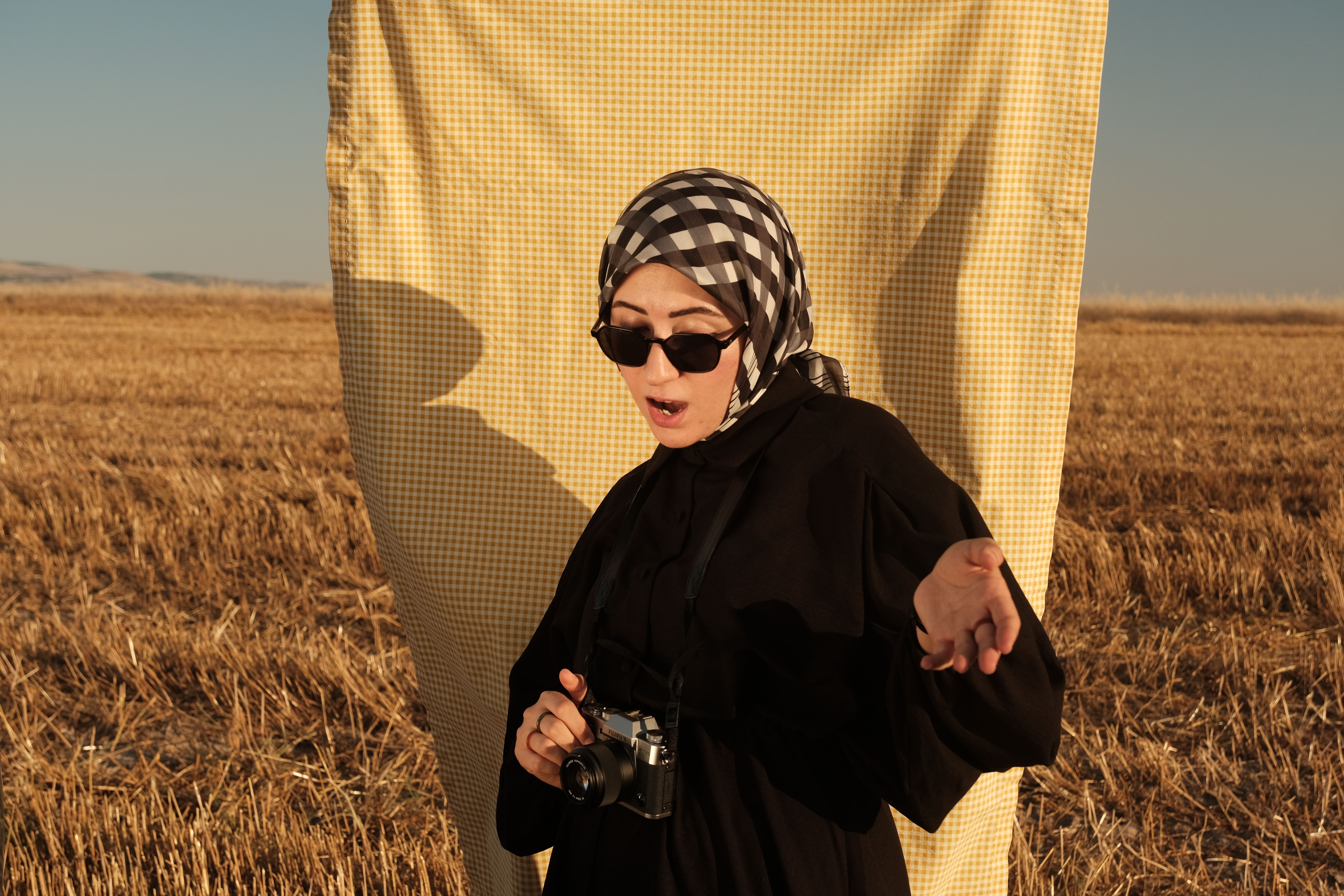 Woman with camera and hijab standing in a sunlit field, expressing surprise.