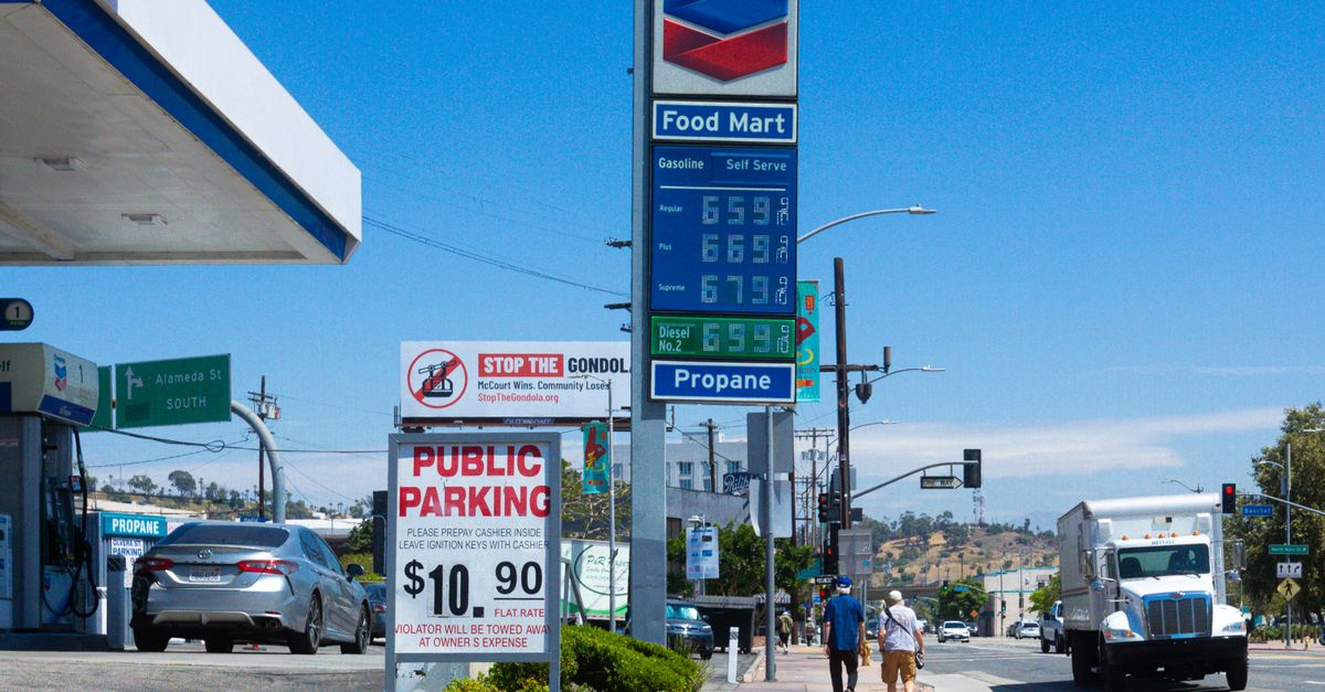 Photo by David Brown People walking near a Chevron gas station with prominent signage and parked vehicles on a sunny day.